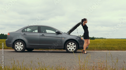 A young girl in a short dress near a broken car is trying to stop a car. The car suddenly broke down on a country road down while driving.		
