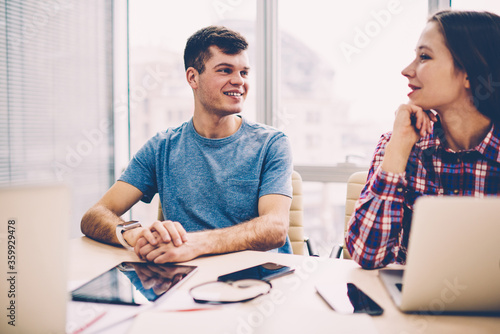 Cheerful male and female colleagues dressed in casual wear laughing during discussion of creative ideas sitting at meeting table in office.Positive employees talking with each other about project