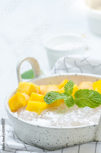 The thai dessert food modern style : The homemade sago in coconut milk with topping is mango cut into squares and cantaloupe ball in the white bowl and placed on tablecloth plaid on the marble table.