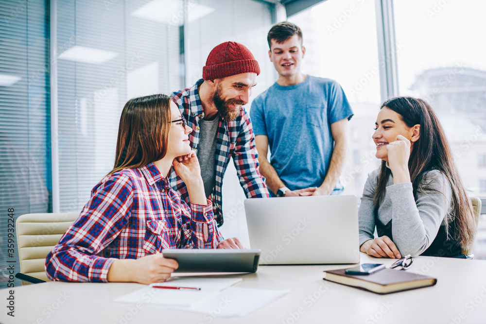 Fototapeta premium Successful group of young professionals in casual wear laughing during cooperation on design project sitting at laptop in office.Positive students teamworking on common task during brainstorming