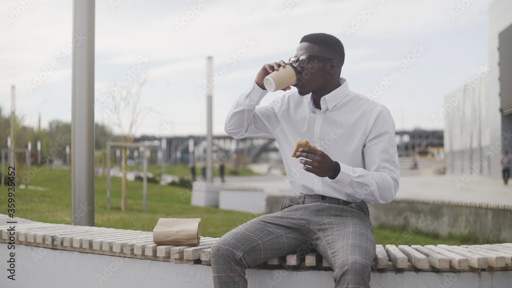 Fototapeta premium Young African American Man drinking coffee or tea and eating sandwitch in park near the office