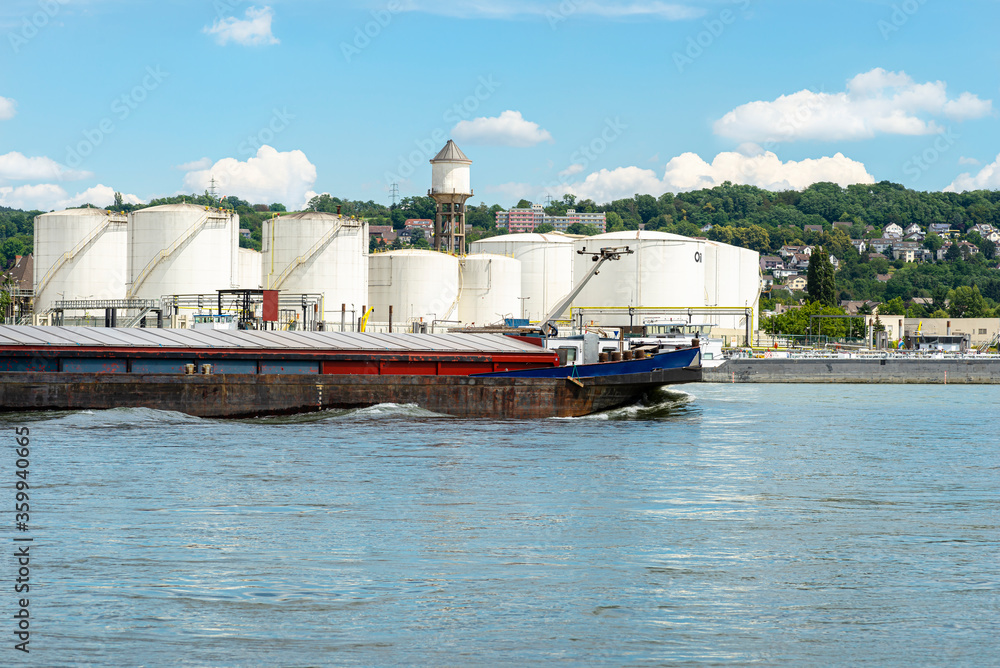 Storage silos, fuel depot of petroleum and gasoline on the banks of the river in western Germany on a beautiful blue sky with clouds. Visible coal barge.