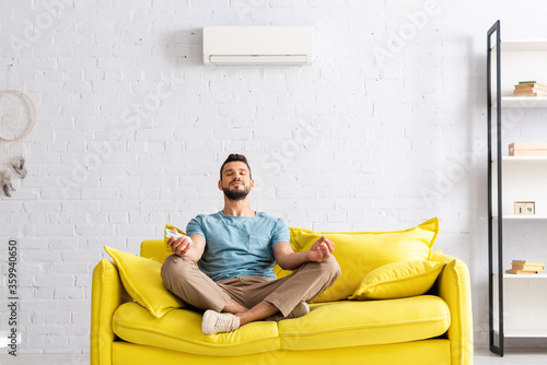 Young man holding remote controller of air conditioner while meditating on sofa