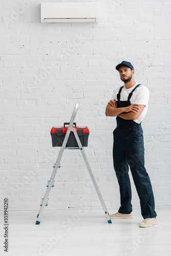 Handsome workman in overalls looking at camera near toolbox on ladder and air conditioner on white wall