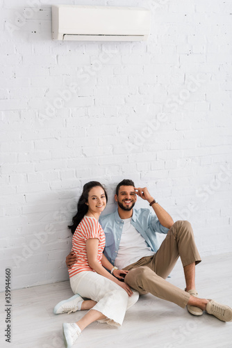 Bearded man embracing girlfriend while sitting on floor near air conditioner on wall at home