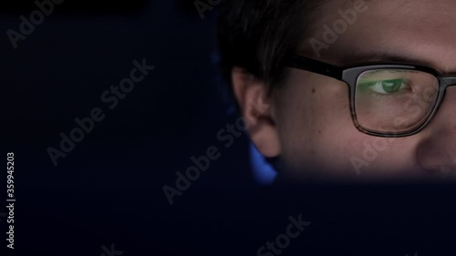 Man in eyeglasses late at night scrolling in front of laptop pan shot left to right. Coder, programmer or developer using laptop in dark. Close up of glasses with reflection of computer screen.