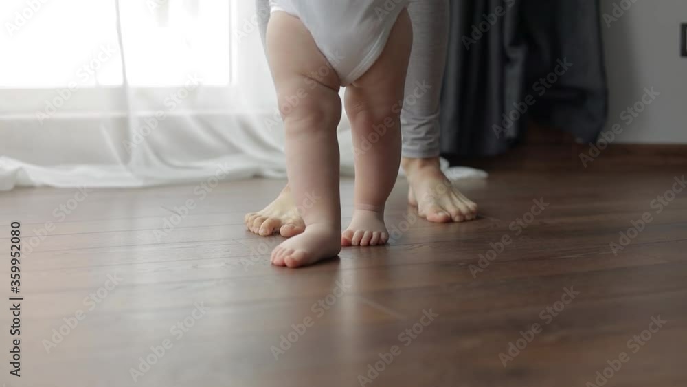 The first steps. A small child learns to walk holding his mother's hands. Caucasian child takes his first steps on the wooden heated warm floor The first steps. A small child learns to walk holding his mother's hands. Caucasian child takes his first steps on the wooden heated warm floor