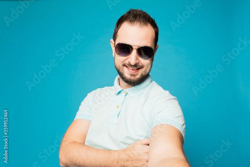 A young bearded man is fooling around at the camera on a blue background. The guy takes a selfie and builds funny faces