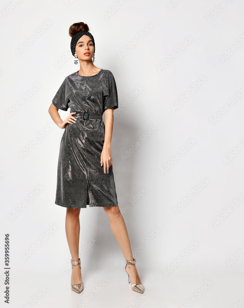 Gorgeous young female with pinned-up hair posing in stylish headband and shiny charcoal dress with belt putting hand on hip, isolated on white Gorgeous young female with pinned-up hair posing in stylish headband and shiny charcoal dress with belt putting hand on hip, isolated on white