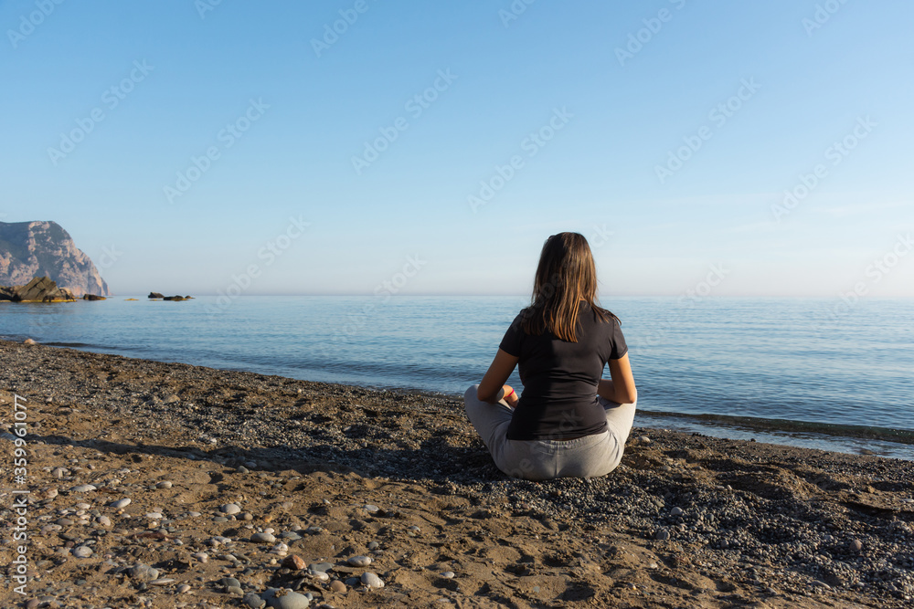 The girl is sitting on the beach. Beautiful brunette girl alone against the background of a blue calm sea. Peace and contemplation. Enjoy the beautiful view. The concept of loneliness. Authenticity.