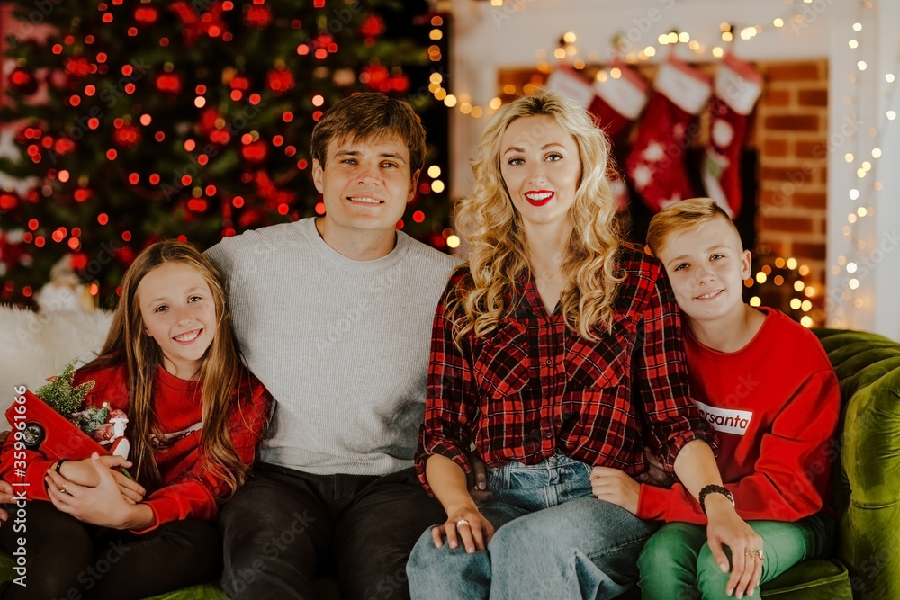 Young happy family of four in Christmas clothes sitting and smiling in living room against Christmas lights. Young happy family of four in Christmas clothes sitting and smiling in living room against Christmas lights.