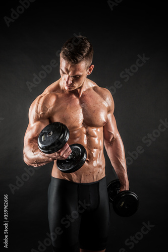 Fitness in gym, sport and healthy lifestyle concept. Handsome athletic man showing his trained body on black background. Bodybuilder male model training biceps muscles with dumbbell.