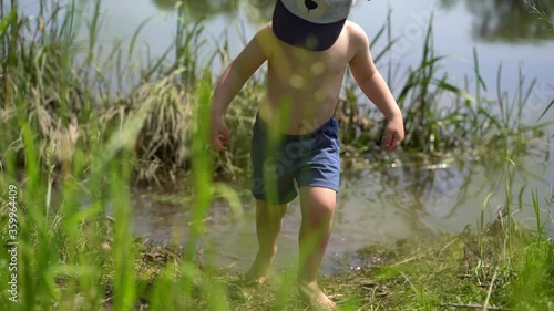 A small boy, a European, dressed in blue shorts and a cap stands with his feet in the river with his back to the camera, turns, gets out of the water and goes to the camera on a Sunny summer day