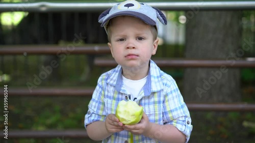 A small boy of European appearance stands at the iron railing in the zoo / Park and eats a green Apple looking at the camera, turns and looks away