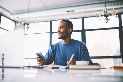 Cheerful afro american male international student doing homework task sitting in university cafeteria with cup of coffee using modern technologies to find information for research from web sources