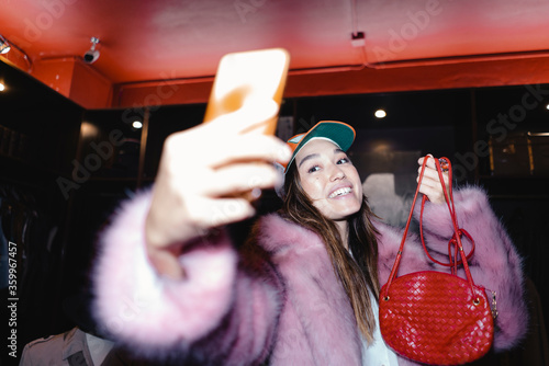 Happy woman taking selfie through smart phone while wearing fur jacket and holding leather purse in store photo