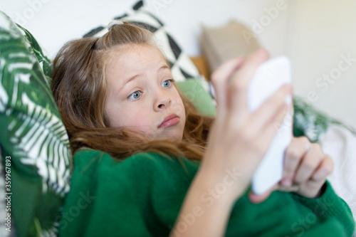 Portrait of girl wearing green pullover lying on bed starring at cell phone
