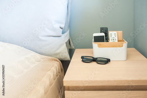 Organizer box for storing personal belongings, stands on the bedside table near the bed. Inside the phone, a remote control for light, near glasses.
