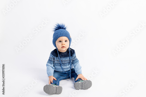 Baby 8 months old boy sitting on a white isolated background in warm winter clothes and a hat, children's fashion, advertising children's clothing, space for text
