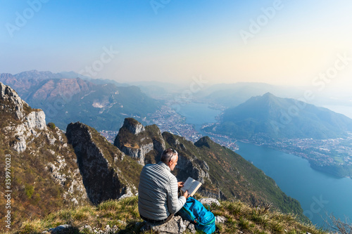 Rear view of hiker reading a book on mountaintop, Orobie Alps, Lecco, Italy