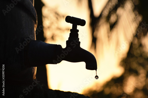 Silhouette of old vintage metal tap leaking water droplet in dry summer morning/evening, water shortage, crisis, scarcity 