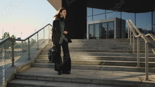 A brunette girl in a black classic suit stands next a business center on the stairs looking around in search of a taxi