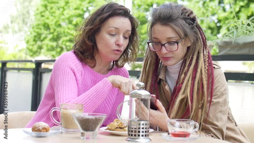 Modern girl with long dreadlocks is showing something on the smartphone to her beautiful female friend in pink sweater during a lunch break together in a coffee shop outdoors Modern girl with long dreadlocks is showing something on the smartphone to her beautiful female friend in pink sweater during a lunch break together in a coffee shop outdoors