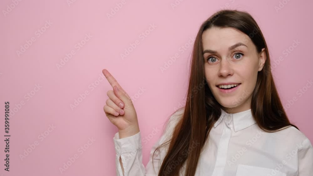 Cheerful excited young woman pointing finger at copy space surprised by unbelievable sale, amazed happy brunette female indicating advertising cheap offer, wears shirt, isolated on pink background Cheerful excited young woman pointing finger at copy space surprised by unbelievable sale, amazed happy brunette female indicating advertising cheap offer, wears shirt, isolated on pink background