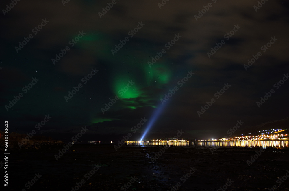aurora borealis over a boat searchlight on the fjord reflecting of the surface with illuminated settlement in the background