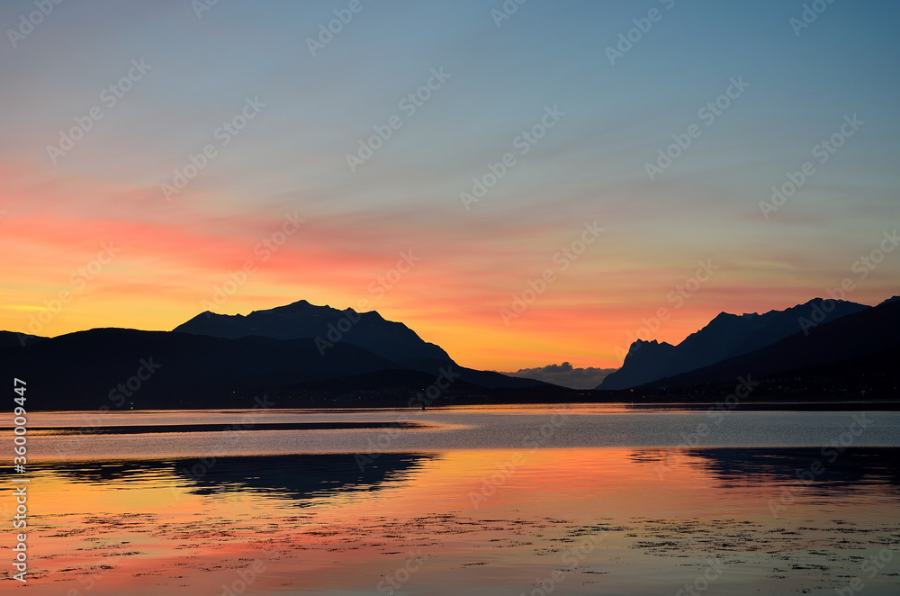 colourful dawn sky behind two majestic mountain with heavy fog coming towards them in the background and colourful reflections on the fjord water in the foreground