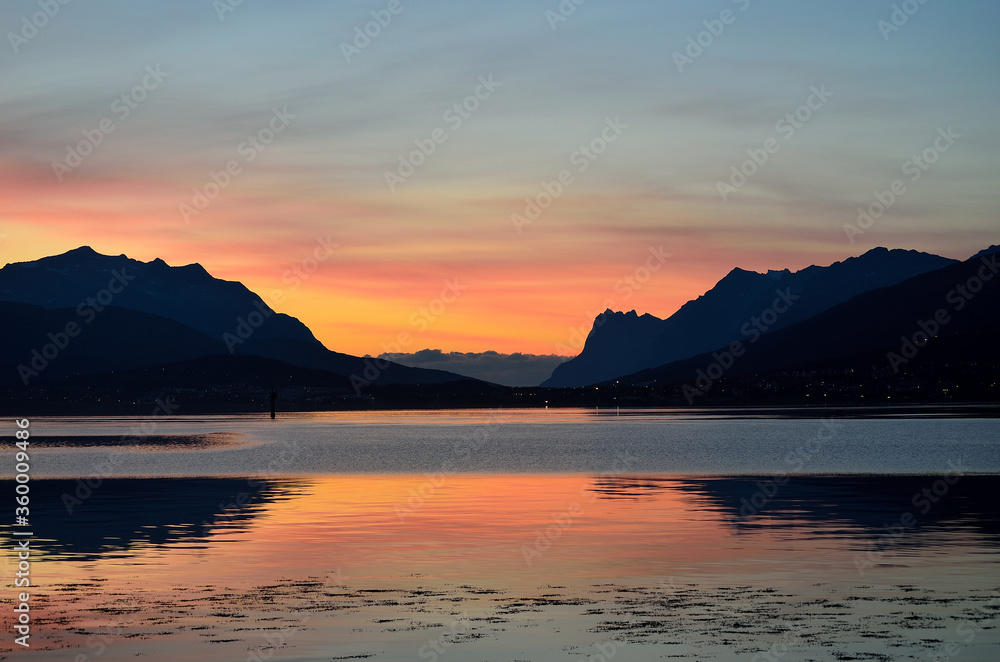 colourful dawn sky behind two majestic mountain with heavy fog coming towards them in the background and colourful reflections on the fjord water in the foreground