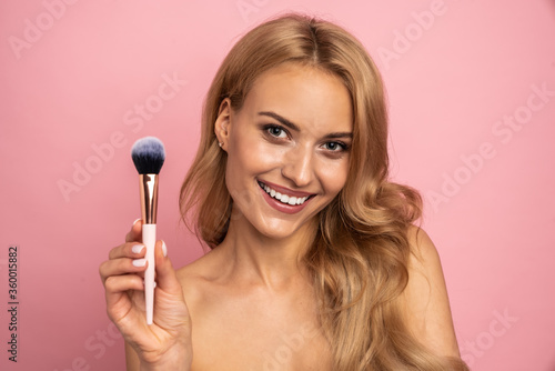 Beauty portrait of a lovely young woman with long blonde hair standing isolated over pink background  holding makeup brush