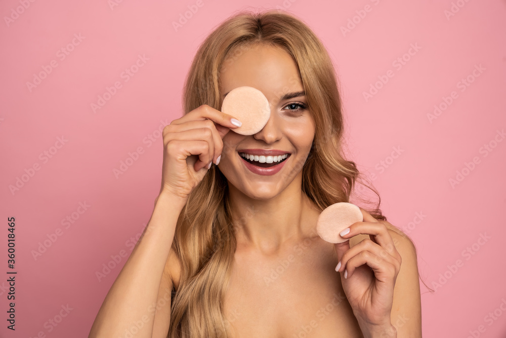 Close up beauty portrait of an attractive smiling young blonde topless woman with bright makeup standing isolated over pink background, holding nakeup sponge Close up beauty portrait of an attractive smiling young blonde topless woman with bright makeup standing isolated over pink background, holding nakeup sponge