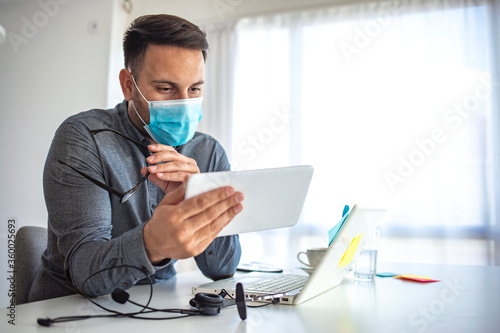 Man in a medical mask at the office. The man stayed at home and works remotely. The guy uses a Tablet PC and a laptop for work, and medicine mask and antiseptic for self protective