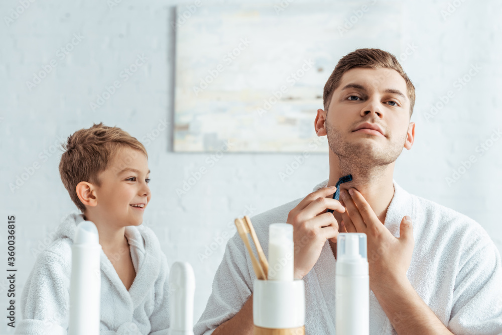 selective focus of young man shaving neck near smiling son in bathroom selective focus of young man shaving neck near smiling son in bathroom