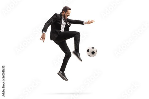 Full length profile shot of a man in a black suit kicking a soccer ball