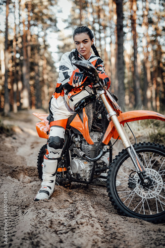 Fototapeta Naklejka Na Ścianę i Meble -  Beautiful young brunette female racer riding motocross bike on a trail of sand in the woods