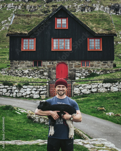 Happy man holding little sheep. Guy on the background of a house with a grass roof in the Faroe Islands. Tourism is an active life and adventure in Scandinavia.