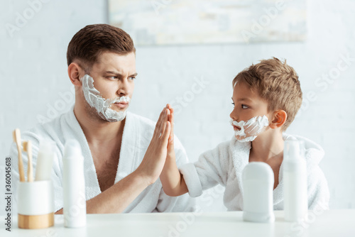 selective focus of serious father and son with shaving foam on faces giving high five