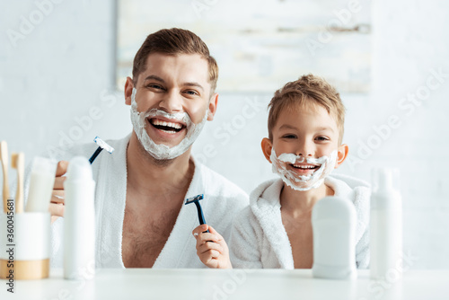 Foto selective focus of cheerful father and son with shaving foam on faces holding sh