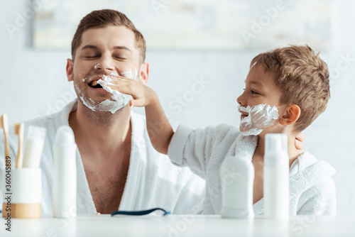 selective focus of smiling boy applying shaving foam on fathers face