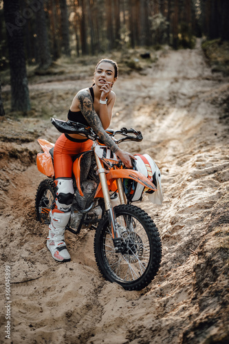 Fototapeta Naklejka Na Ścianę i Meble -  Tattooed female racer wearing motocross outfit with semi naked torso sitting on her bike in the forest