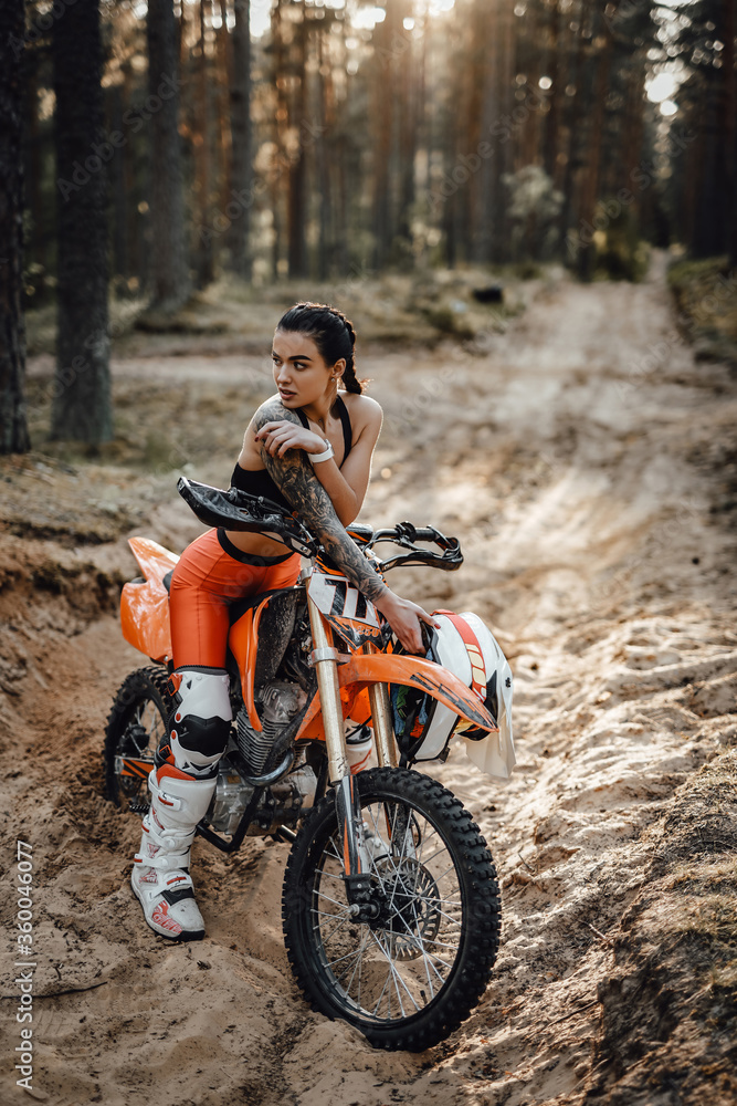 Beautiful female racer wearing motocross outfit with semi naked torso sitting on her bike in the forest
