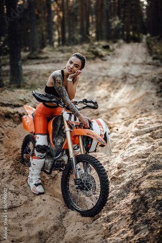 Fototapeta Naklejka Na Ścianę i Meble -  Beautiful female racer wearing motocross outfit with semi naked torso sitting on her bike in the forest