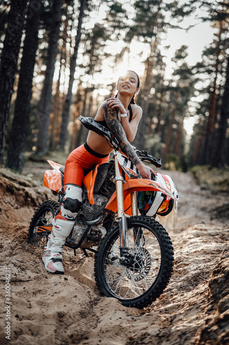 Cheeky tattooed racer girl wearing motocross outfit with semi naked torso sitting on her bike at the bright sunset in the forest