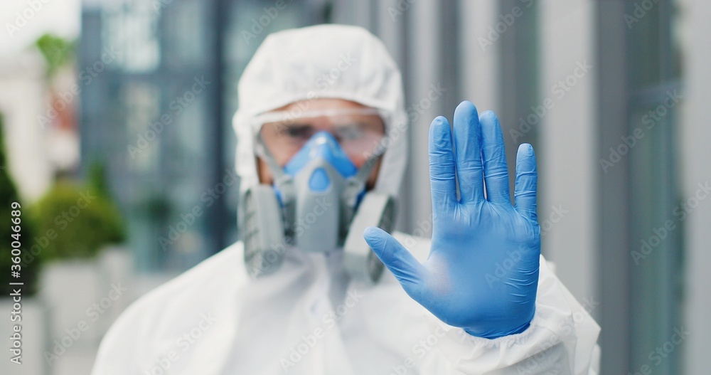 Caucasian man in white suit, goggles, gloves and respirator looking at camera and showing gesture stop with hand. Male healthcare worker of disinfection prohibitting pass outdoor. Prohibition to enter Caucasian man in white suit, goggles, gloves and respirator looking at camera and showing gesture stop with hand. Male healthcare worker of disinfection prohibitting pass outdoor. Prohibition to enter