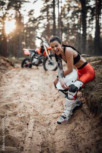 Beautiful brunette girl wearing motocross outfit with semi naked torso sitting on grass and unrestrained laughs