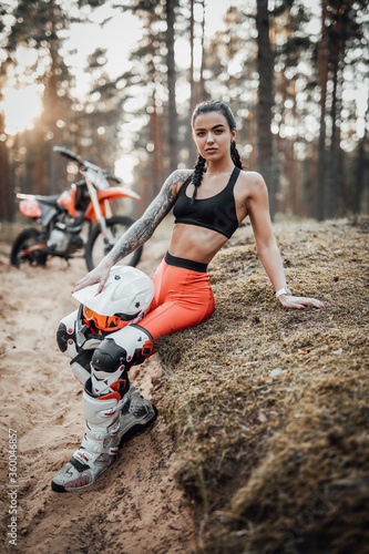 Beautiful brunette girl wearing motocross outfit with semi naked torso sitting on a grass in the forest
