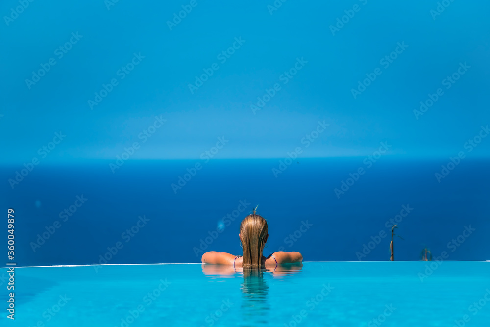 Summer lifestyle in an infinity pool with a young blonde Caucasian woman in a pink and purple bikini with sunglasses. Leaning against the bottom of the pool looking at the sea and the blue sky