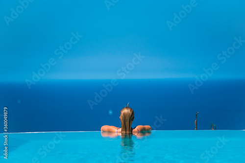 Summer lifestyle in an infinity pool with a young blonde Caucasian woman in a pink and purple bikini with sunglasses. Leaning against the bottom of the pool looking at the sea and the blue sky